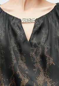 Schwarze Bluse mit strukturiertem Schlangenmuster, ausgestattet mit einem V-Ausschnitt und einer goldenen dekorativen Schnalle am Kragen.