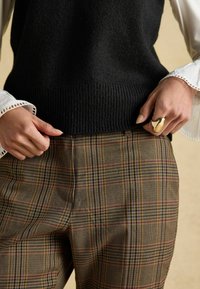 Schwarzes gestricktes ärmelloses Oberteil mit geripptem Saum, kombiniert mit braunen Karo-Hosen mit beige, blau und roten Streifen; goldener Ring am Handgelenk.