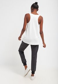Weißes ärmelloses Tanktop kombiniert mit schwarzen Jogginghosen mit elastischen Bündchen. Das Model steht mit dem Rücken zur Kamera und trägt weiße Sneaker.