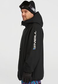 Zwarte waterdichte jas met een capuchon, met een logo op de mouw en handschoenen. Heldergekleurde broek met patroon is zichtbaar onder de jas.