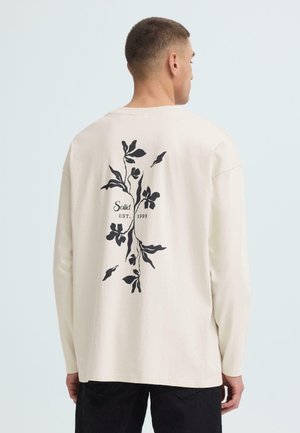 Mann trägt cremefarbenes Langarmshirt mit schwarzem Blumenmuster und Aufschrift "Solid EST. 1999" auf dem Rücken, steht vor hellem Hintergrund.