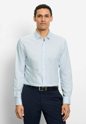 Mann trägt hellblau gemustertes Hemd mit langen Ärmeln, in dunkelblaue Hose mit schwarzem Gürtel gesteckt, steht vor schlichter weißer Wand.