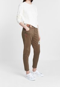 Hnědé slim-fit kalhoty zdobené květinovou výšivkou, zipovou kapsou a dekorativním šitím. V kombinaci s bílým texturovaným svetr a bílými teniskami.