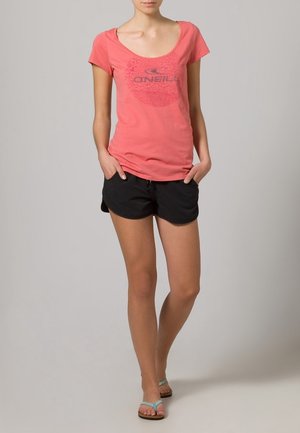 Femme portant un t-shirt corail O'Neill, un short noir et des tongs bleues, debout les mains dans les poches contre un fond uni.