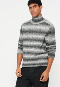 Szary sweter z golfem, z gradientowym wzorem pasków, ściągaczami przy mankietach i miękką teksturą. Połączony z ciemnymi dżinsami, noszony wygodnie.