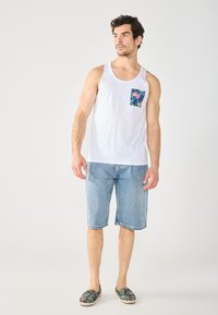 White tank top with a blue floral pocket, light blue denim shorts, and patterned slip-on shoes, standing on a plain background.