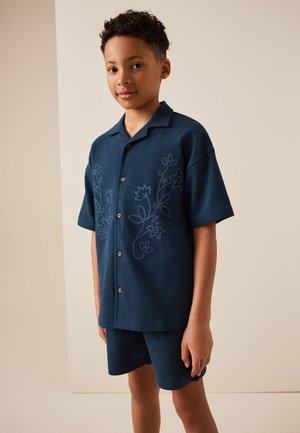 Dreng står iført en mørkeblå kortærmet skjorte med blomsterbroderi og matchende shorts mod en ensfarvet beige baggrund.