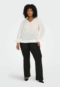 Bijela, prozračna bluzica s cvjetnim uzorkom i V-izrezom; napuhane duge rukave s teksturiranim manžetama. Uparen s crnim hlačama i potpeticama.