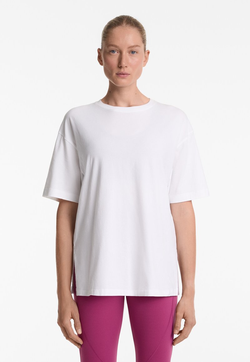 Hvid oversized t-shirt i blød bomuld med rund halsudskæring, korte ærmer og slidser i siderne ved kanten. Simpelt design og glat tekstur.