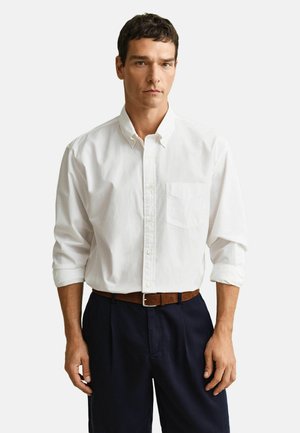 Mann trägt ein weißes Hemd mit hochgekrempelten Ärmeln, dunkelblaue Hose und einen braunen Gürtel, steht vor einem schlichten Hintergrund.