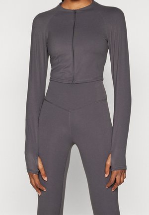 Kvinde iført mørkegrå crop top med lange ærmer og lynlås foran samt matchende høj taljede leggings med hul til tommelfingrene i ærmerne.