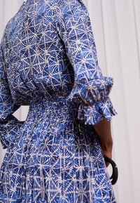 Vestido azul con un patrón de espirales geométricas blancas, con una cintura fruncida, mangas acampanadas y un bolso negro en forma de círculo.