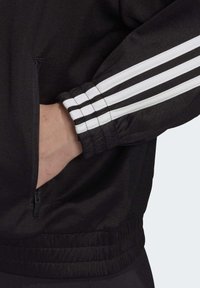 Black athletic jacket with white stripes on the sleeve, featuring a zip pocket and elastic cuffs, made of smooth synthetic material.