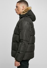 Giacca puffa nera con cappuccio, caratterizzata da un bordino in pelliccia sintetica e motivi con logo in rilievo. Ha un design trapuntato e tasche con zip.
