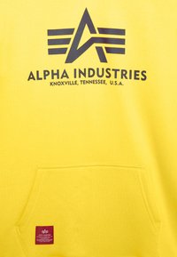 Sudadera amarilla con un bolsillo frontal. Presenta un logo negro que dice "ALPHA INDUSTRIES" y diseños geométricos arriba. Tela de algodón texturizada.