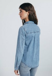 Lichtblauwe denim shirt met een knoopsluiting kraag, lange mouwen en een relaxte pasvorm. Heeft een achterkant yoke en zijsplitten aan de zoom.