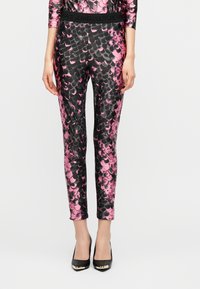 Γυναίκα ντυμένη με ροζ και μαύρο κολάν με μοτίβο λεπιών και αντίστοιχο τοπ, φορώντας μαύρα ψηλά τακούνια, σε απλό φόντο.