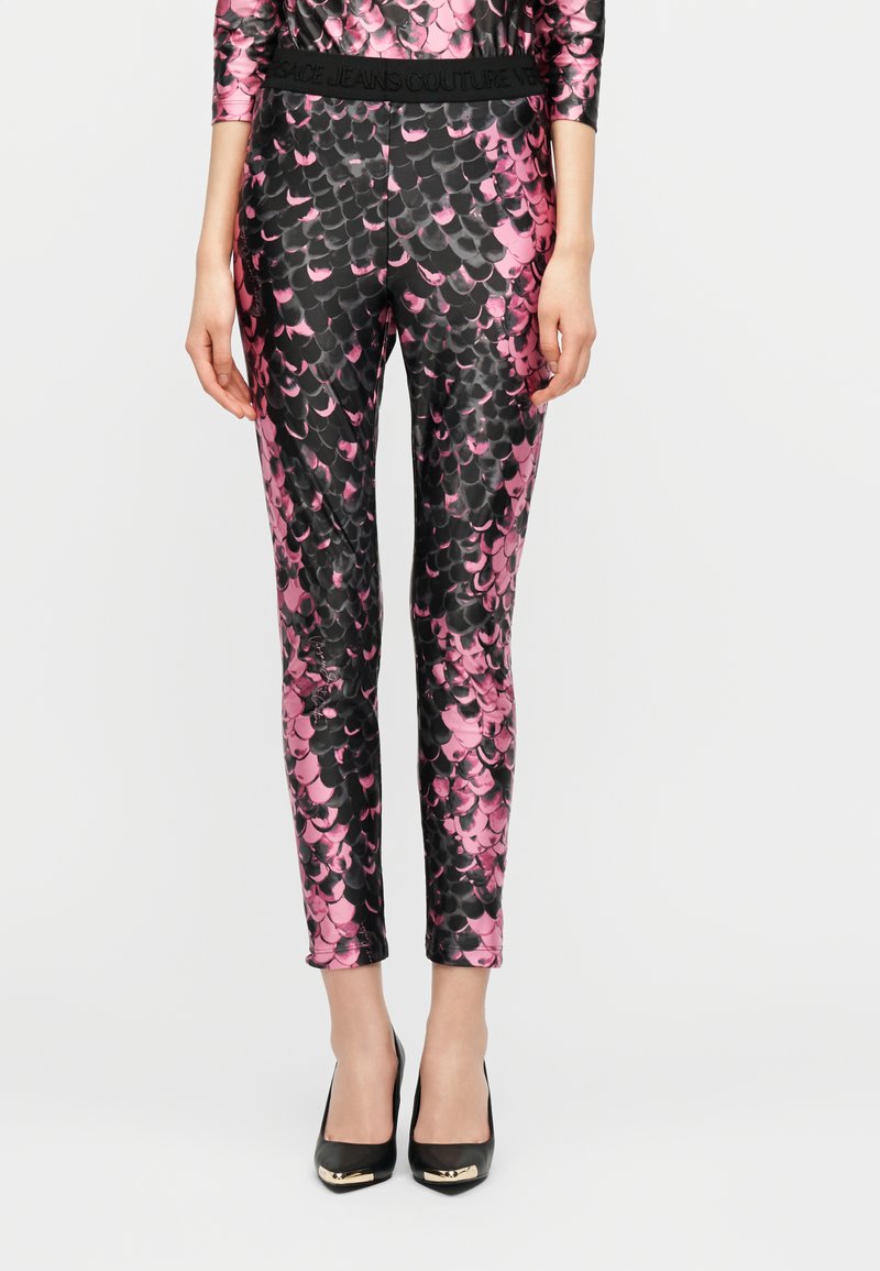 Γυναίκα ντυμένη με ροζ και μαύρο κολάν με μοτίβο λεπιών και αντίστοιχο τοπ, φορώντας μαύρα ψηλά τακούνια, σε απλό φόντο.