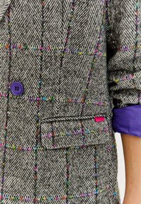Šedý blazér v herringbone vzoru s vícebarevným sekvencovým mřížkovým vzorem, fialovým knoflíkem, klopovou kapsou s růžovou cedulkou a ohrnutými fialovými manžetami.