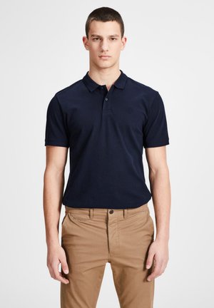 Polo-shirt bleu marine en coton, avec un col classique, des manches courtes et un logo discret sur la poitrine gauche, associé à un pantalon beige.