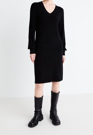 Personne portant une robe noire à manches longues, col en V, longueur genou, associée à des bottes en cuir noires mi-mollet, debout devant un fond uni.