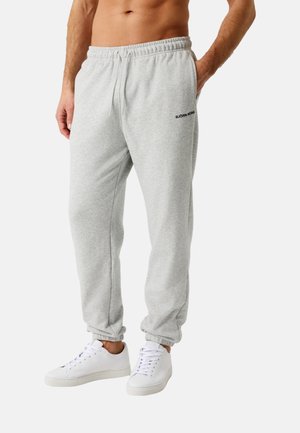 Egy férfi világosszürke Björn Borg melegítőnadrágot és fehér tornacipőt visel, egyik kezét a zsebében tartva, egyszínű fehér háttér előtt áll.