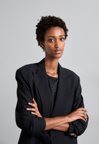 Zwarte blazer met een op maat gemaakt ontwerp, brede revers en zilveren knoopdetails. Draagt over een zwart shirt met een gladde textuur en pasvorm.