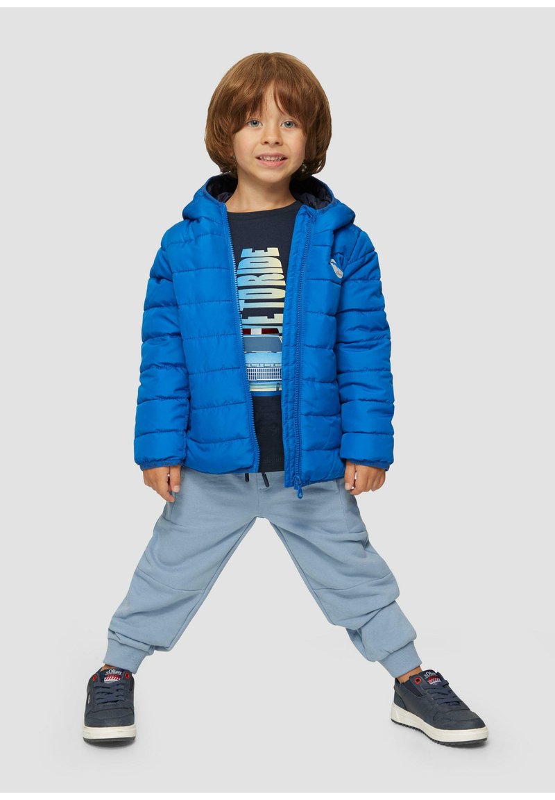 Blaues Puffer-Jacke mit Kapuze, leichtes Design, gesteppte Textur. Dunkelgraues T-Shirt darunter mit Grafikdruck. Hellblaue Jogginghose. Schwarze Sneakers.