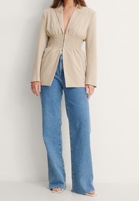 Beżowa marynarka z marszczonym pasem, długimi rękawami i szpiczastymi klapami, dopasowana do jasnoniebieskich spodni szerokich nogawkach i neutralnych sandałach.