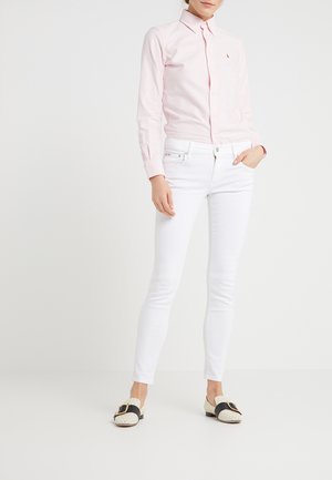 Femme portant une chemise à rayures rose clair avec des boutons, un pantalon slim blanc et des mocassins blancs avec des brides noires et de grandes boucles.