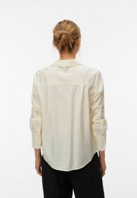 Chemise légère crème à manches longues, avec des accents décoratifs en dentelle sur les coudes et un ourlet légèrement arrondi à l'arrière.