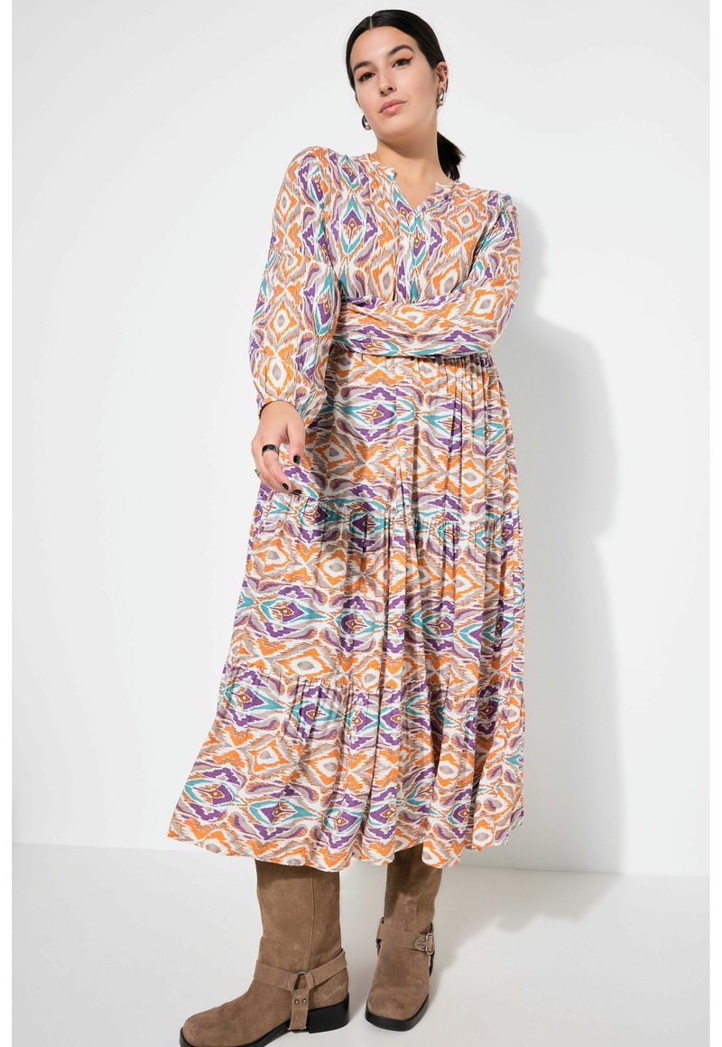 Abito maxi fluido in vivace arancione, viola e verde con motivo geometrico. Presenta scollo a V, maniche lunghe e vita elastica. Abbinato a stivali marroni.