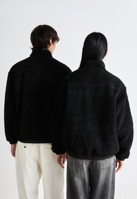 Black fleece jackets with high collars, featuring embossed "TOMMY JEANS" text. Elastic hems, textured fabric, and relaxed fit.