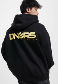 Svart hoodie med guld "DNGRS INVADER" text på ryggen. Har en rymlig design, justerbar huva och ribbade ärmslut. Mjuk tygkänsla.