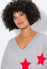 Maglione lavorato a maglia grigio chiaro con scollo a V, decorato da due evidenti applicazioni a forma di stella rossa sul davanti. Texture morbida e vestibilità comoda.