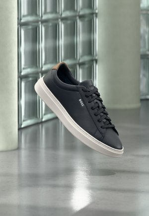 Czarny niskoprofilowy sneaker sznurowany z białą podeszwą i brązową zakładką na pięcie, markowany jako „BOSS”, unoszący się w pomieszczeniu obok ściany z bloków szklanych.