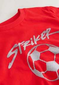 T-shirt en coton rouge avec texte "Striker" argenté texturé et motif de ballon de football, avec encolure côtelée et texture de tissu lisse.