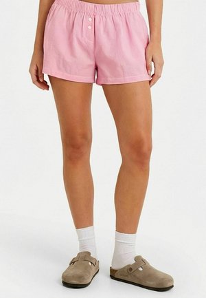 Vrouw die lichtroze elastische shorts met twee knopen draagt, witte sokken en taupekleurige instappers, staande op een witte achtergrond.