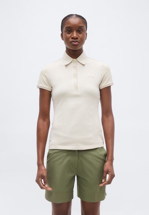 Γυναίκα στέκεται με το πρόσωπο προς τα εμπρός φορώντας μια μπεζ πόλο μπλούζα με κοντά μανίκια και χακί σορτς, μπροστά σε απλό ανοιχτό φόντο.