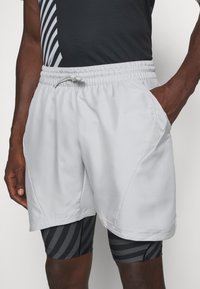 Pantalones cortos deportivos de color gris claro con una cinturilla elástica, bolsillos laterales y paneles de malla en contraste. Llevados sobre pantalones cortos de compresión a rayas negras.