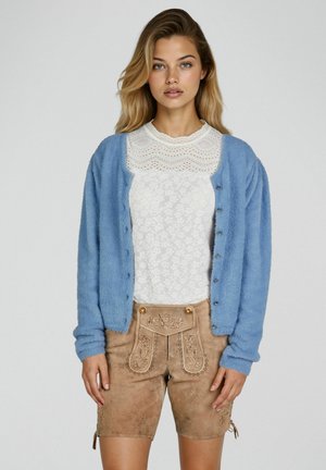 Junge Frau trägt eine blaue, flauschige Strickjacke über einem weißen Spitzenoberteil und beige bestickte Leder-Shorts, stehend vor einem einfarbigen Hintergrund.