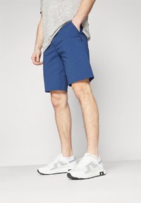 Men's blue shorts made of lightweight fabric, featuring side pockets and a smooth texture. Paired with white sneakers and a gray striped shirt.