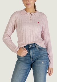 Maglione rosa chiaro lavorato a maglia con motivo a trecce, colletto, tre bottoni e piccolo dettaglio a cuore rosso, abbinato a jeans di jeans blu a vita alta.