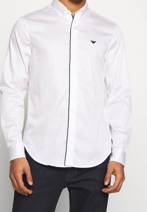 Homme portant une chemise blanche ajustée à manches longues avec une finition noire verticale et un jean foncé, debout devant un fond uni.