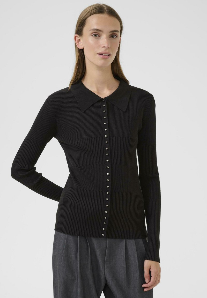 Mujer vistiendo un cardigan ajustado negro de fondo acanalado con un collar ancho y pantalones de sarga grises de talle alto y con pliegues, de pie frente a un fondo liso.