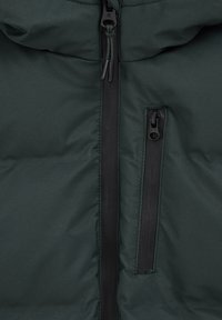 Puhasta jakna v temno zeleni tkanini z osrednjim zadrgo, dvema žepoma z zadrgo in prešito teksturo. Črni detajli na zadrgah.