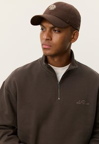 Καφέ φούτερ με φερμουάρ από μαλακό ύφασμα με υφή, με ψηλό γιακά και διακριτική κεντημένη λεπτομέρεια λογότυπου. Περιλαμβάνεται καφέ καπέλο.