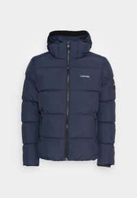 La doudoune bleu marine présente un col montant avec capuche, une fermeture éclair frontale et des poches latérales. Logo imprimé sur la poitrine gauche et la manche.