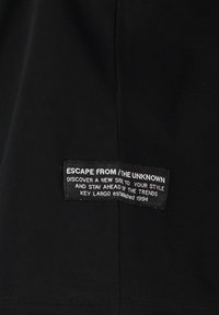 Tela negra con textura acanalada que presenta una etiqueta rectangular con texto blanco. La etiqueta incluye el nombre de la marca y múltiples frases de estilo.