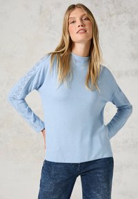 Hellblauer Pullover mit langen Ärmeln, der über einen hohen Kragen und dekorative Stickereien entlang der Ärmel verfügt. Weicher Stoff, lässige Passform.